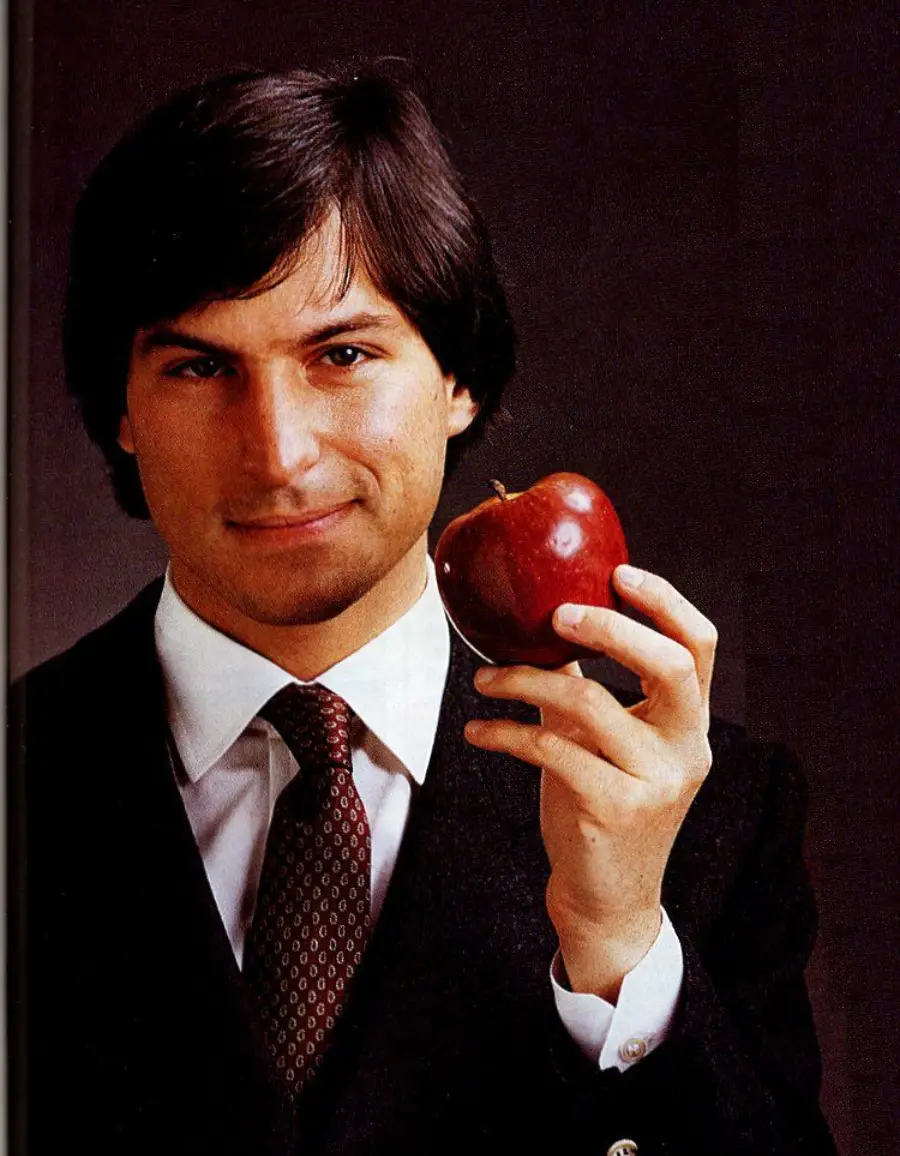 A man in a suit holds up a red apple while looking at the camera, standing against a dark background.