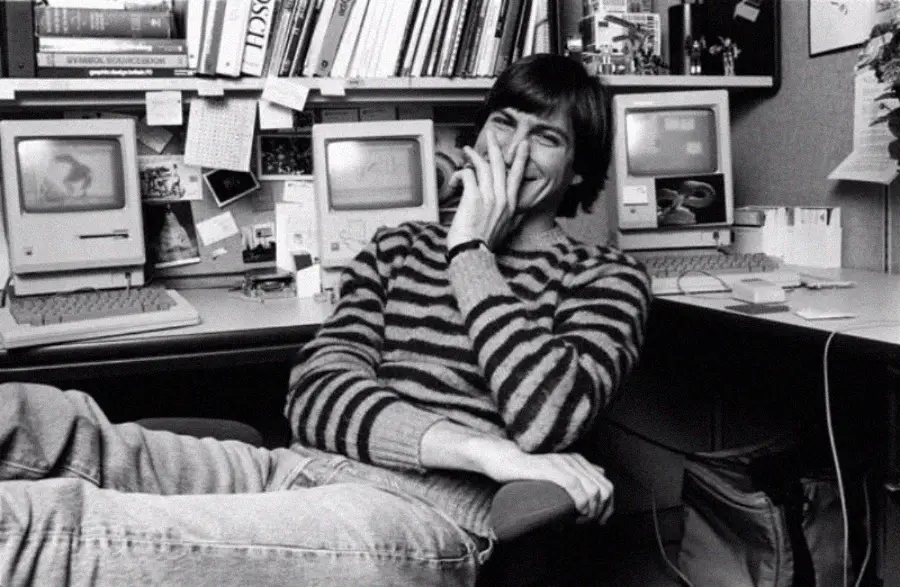 A young man in a striped sweater and jeans sits relaxed in an office chair, smiling and covering his mouth with one hand. Behind him are shelves with books and two vintage Macintosh computers.