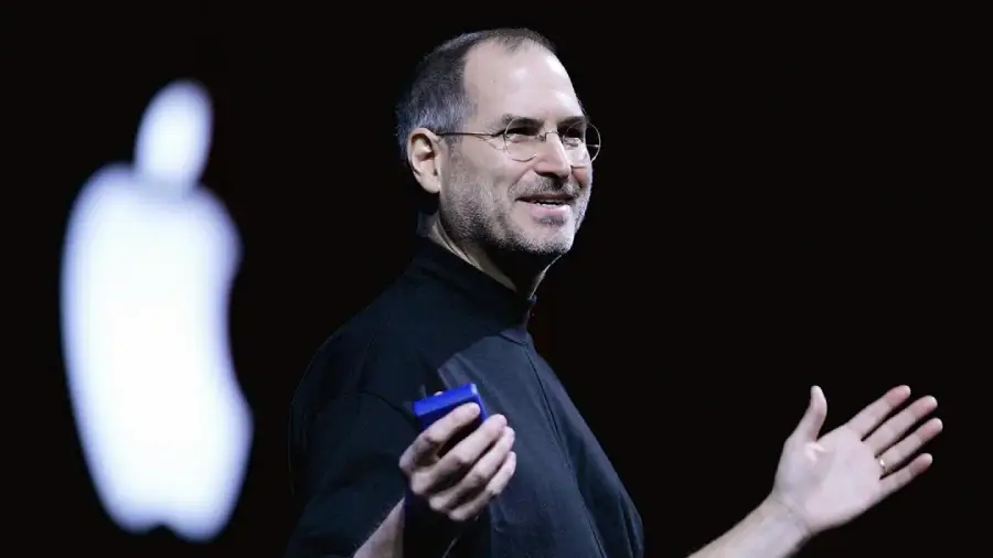 A man in glasses and a black turtleneck speaks on stage, holding a small device, with a blurred white Apple logo visible in the background.
