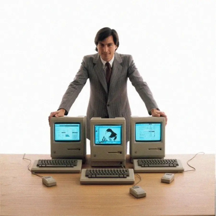 A man in a suit stands behind a wooden desk with three vintage Macintosh computers, each with a mouse and keyboard, against a plain white background.