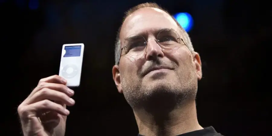 A person with glasses holds up a small white portable media player, smiling slightly, against a dark background.