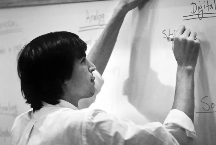 A person in a white shirt writes with a marker on a whiteboard, with the words "Analog" and "Digital" visible at the top. The image is in black and white.