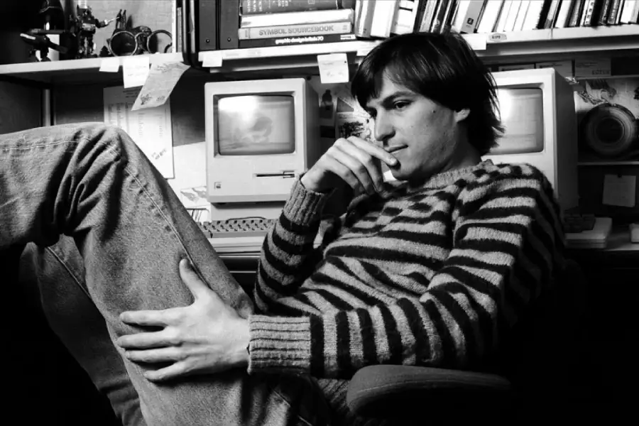 A young man in a striped sweater sits in a chair with his legs up, looking thoughtful. He is in front of an old computer in a cluttered office with books and notes on the shelves behind him. Black and white photo.