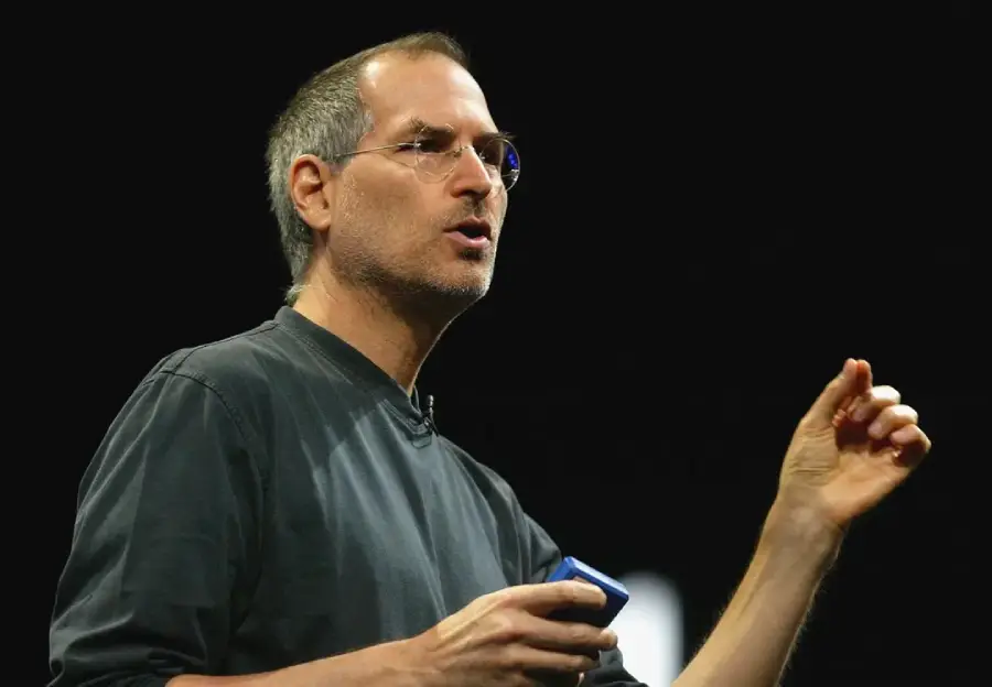 A man wearing glasses and a black long-sleeve shirt is speaking on stage, gesturing with one hand and holding a small object in the other, against a dark background.
