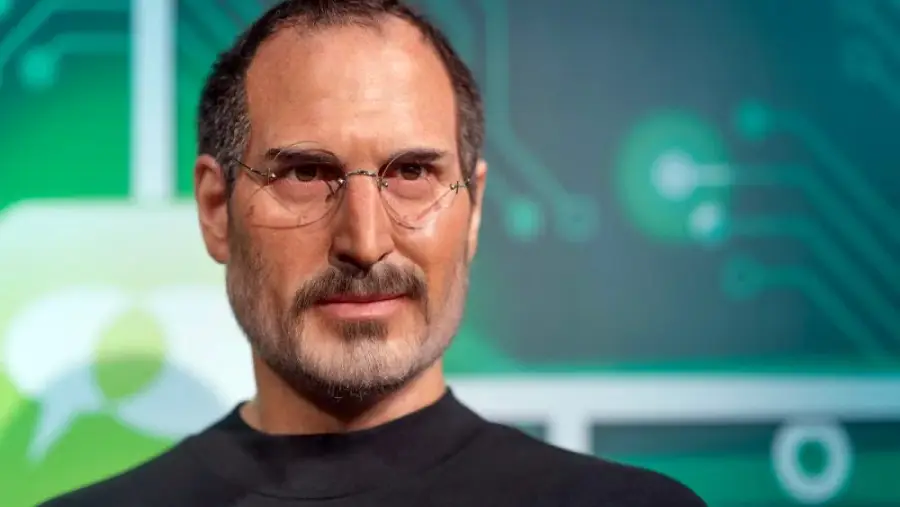 A realistic wax figure of a man with short dark hair, wire-framed glasses, a trimmed beard, and a black turtleneck, set against a green and blue background with circuit-like designs.