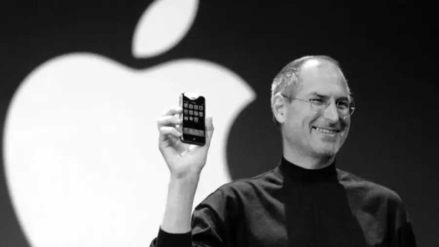A man wearing glasses and a black turtleneck holds up a smartphone while smiling, standing in front of a large Apple logo.