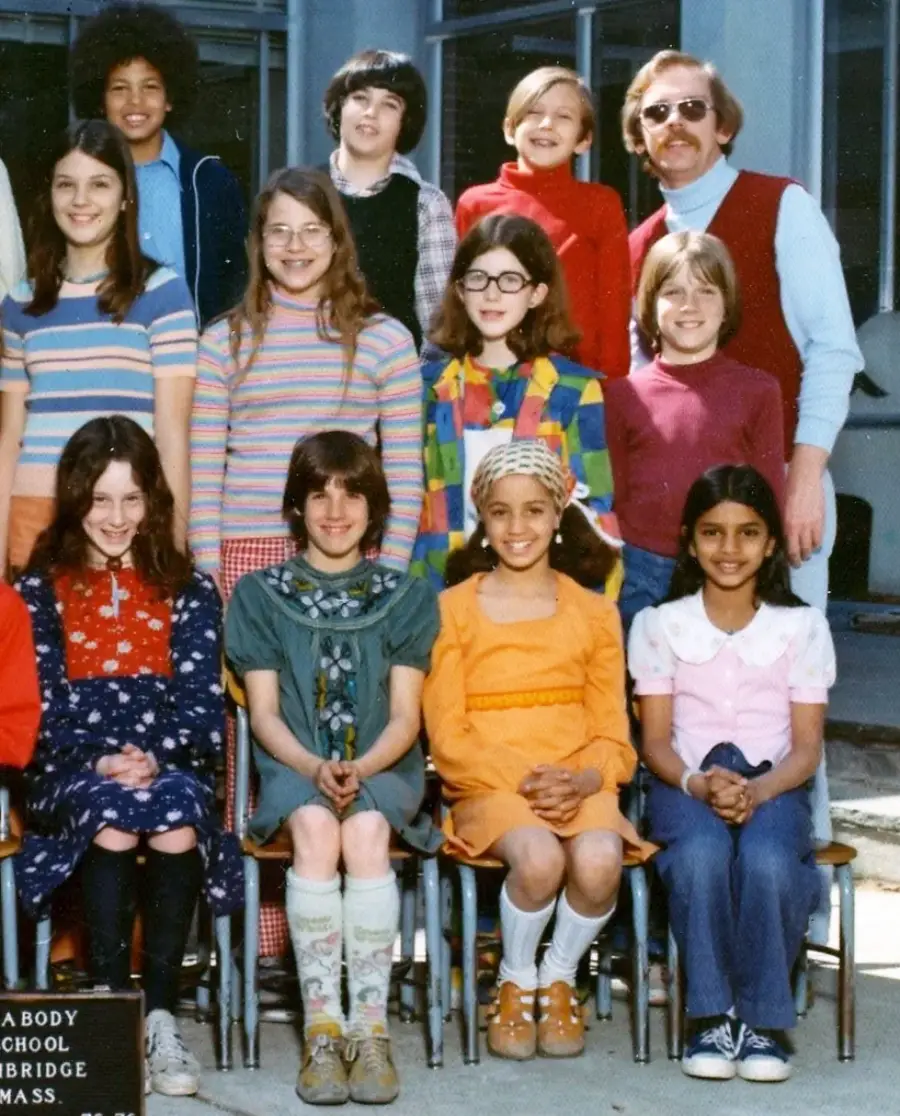 A group of schoolchildren and a male teacher pose outdoors for a class photo. The children wear colorful 1970s-style clothing and are seated or standing in rows, smiling at the camera.
