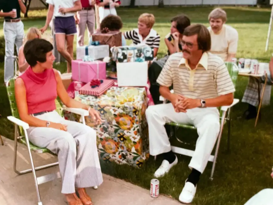 A group of people sit outdoors on lawn chairs around a table with wrapped gifts. Two people are seated in front, talking, while others chat and watch in the background. It appears to be a casual gathering or party.