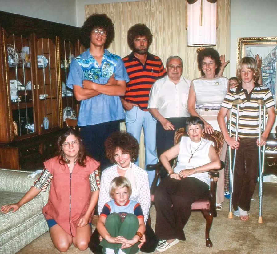A group of ten people, including adults and children, pose in a living room. One child is on crutches, and another sits cross-legged on the floor. The scene has a vintage feel with 1970s-style clothing and decor.