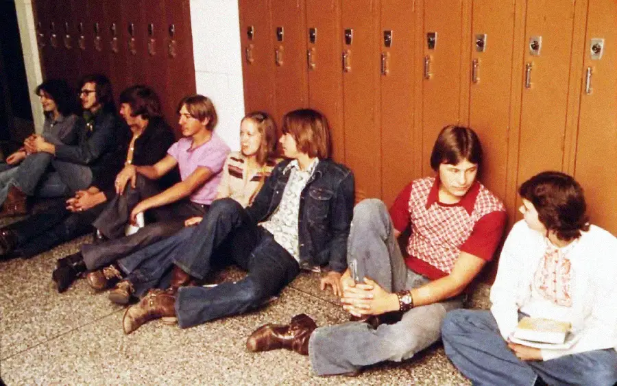 A group of teenagers in 1970s-style clothing sit on the floor in a school hallway, leaning against orange lockers and talking in pairs or small groups.