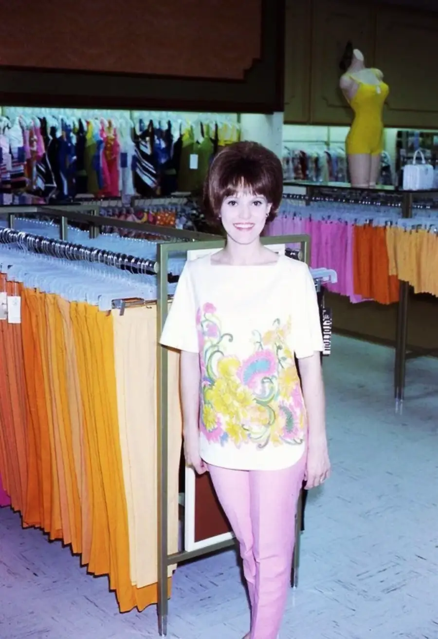 A woman with short brown hair, wearing a white floral top and pink pants, stands and smiles in a clothing store with colorful skirts or pants hanging on racks and swimsuits displayed in the background.