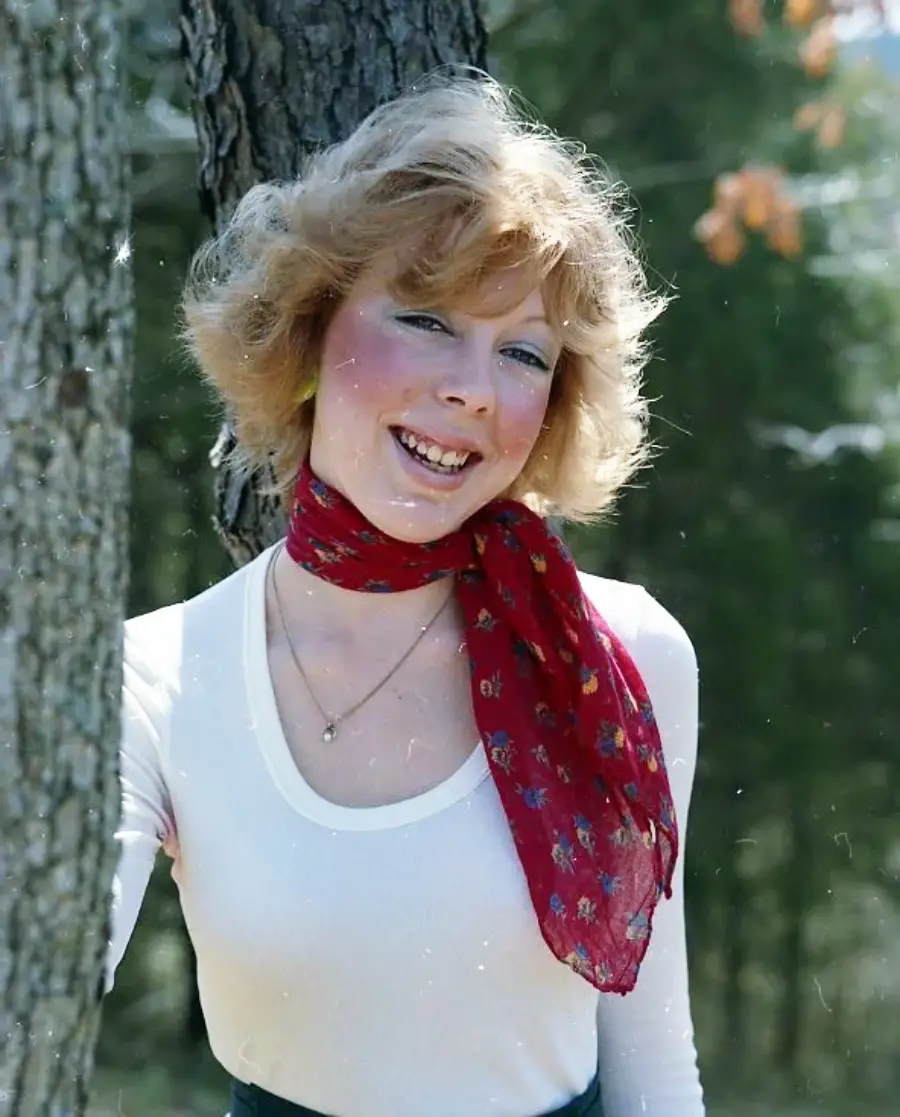 A woman with short blonde hair, wearing a white top and a red patterned scarf around her neck, smiles while standing outdoors next to a tree. The background is filled with greenery.