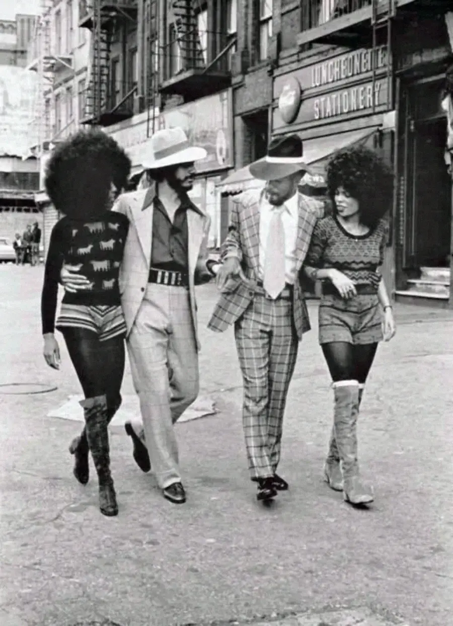 Four stylish people with large afros, two men in patterned suits and hats, and two women in patterned tops, shorts, and high boots, walk arm-in-arm down a city street in a vintage black-and-white photo.