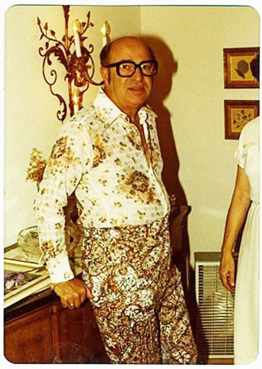 A man wearing large glasses, a floral-patterned shirt, and paisley-print pants stands indoors, leaning on a piece of furniture. Part of another person in a white dress is visible beside him.