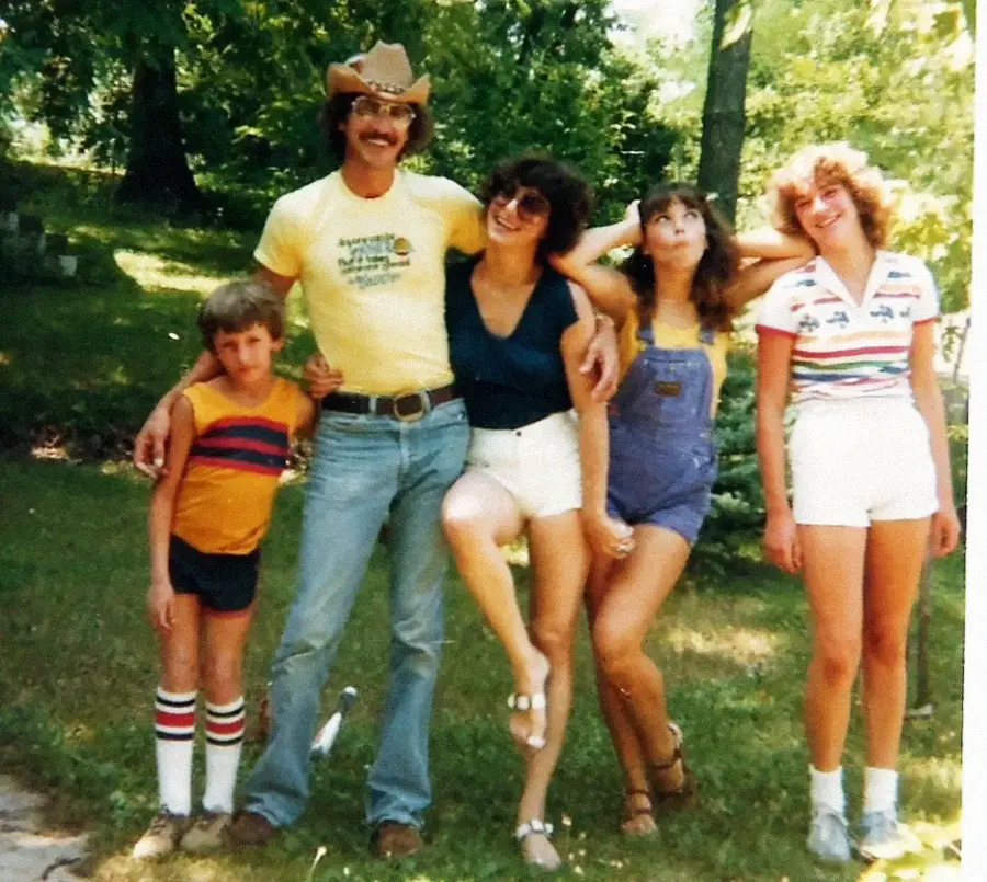 A man in a cowboy hat poses outdoors with four smiling children, all dressed in retro summer clothes. They stand on grass with trees in the background, exuding a joyful, carefree 1970s vibe.