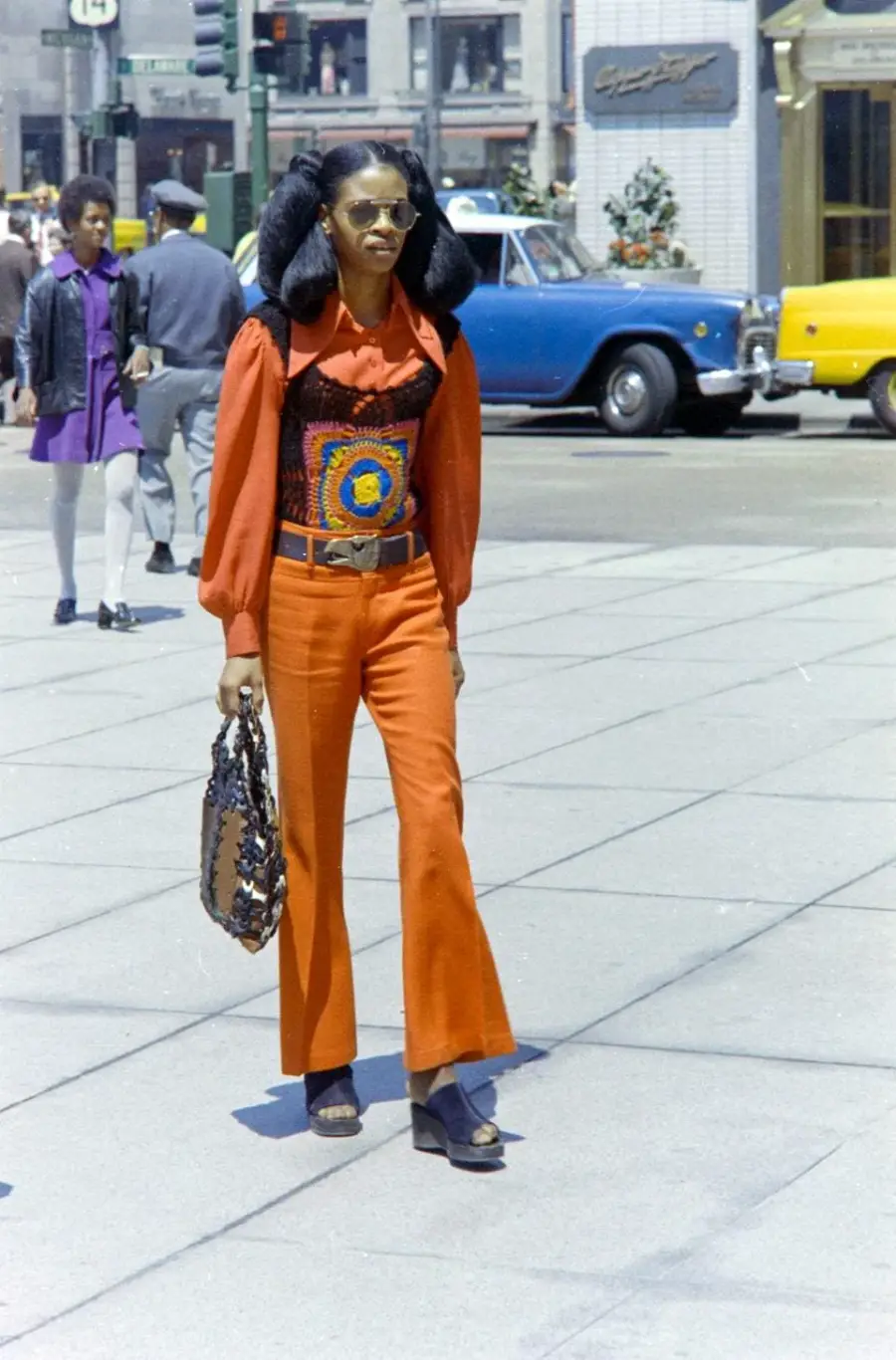A stylish woman in orange flared pants, a bright blouse, and sunglasses walks on a city street holding a large handbag. Vintage cars and pedestrians are visible in the background.