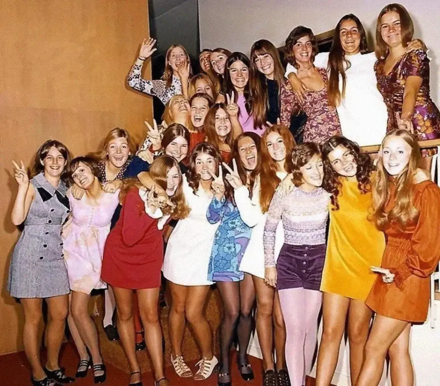 A group of young women in colorful 1970s-style dresses and skirts pose together indoors, many smiling and flashing peace signs. They stand on and around a staircase, celebrating and enjoying themselves.