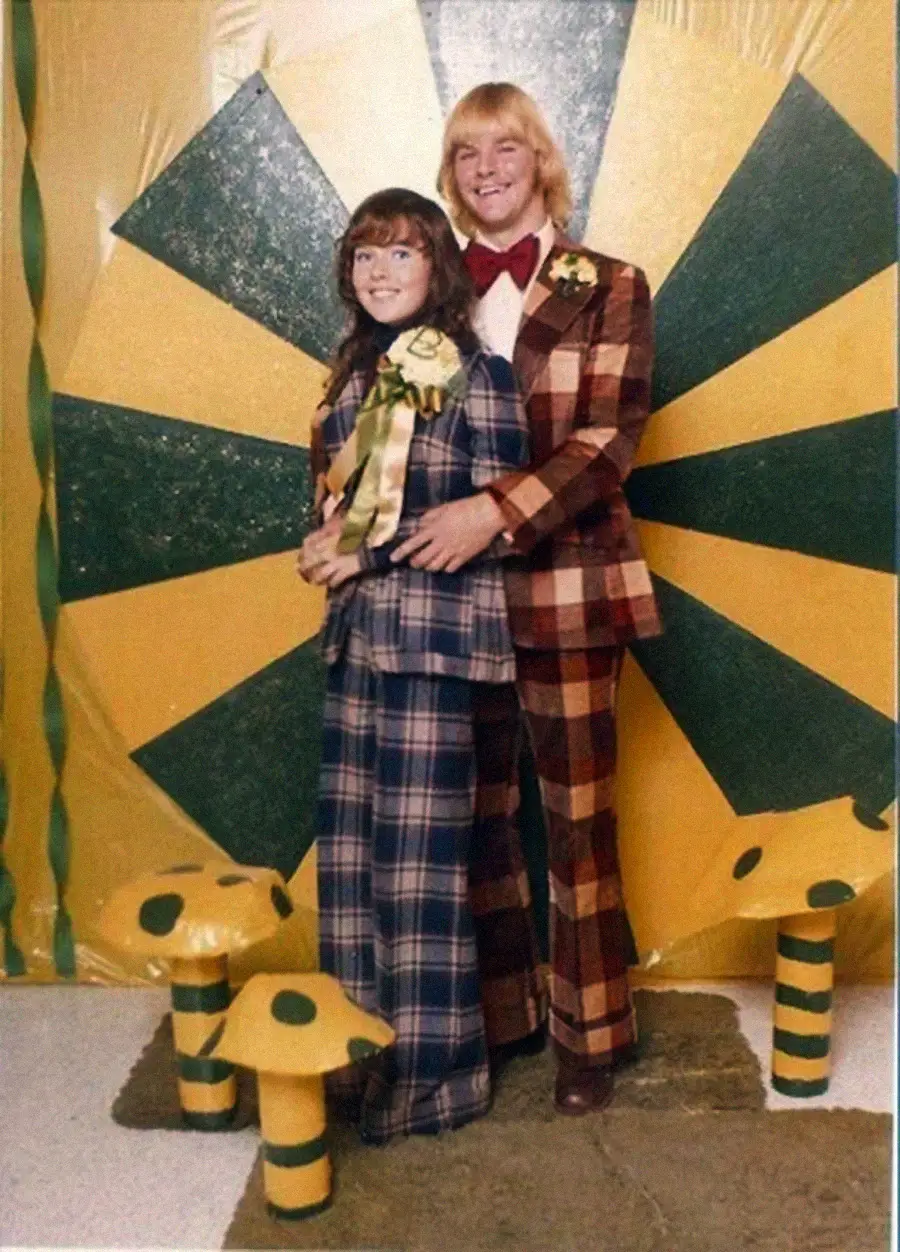 A smiling couple in plaid suits poses in front of a yellow and green sunburst backdrop with matching mushrooms on the floor, evoking a retro or 1970s prom theme.