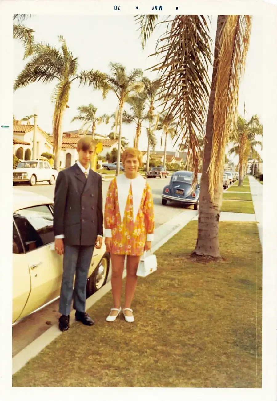 A young man in a suit and a young woman in a colorful coat dress stand on a palm-lined suburban sidewalk next to a white car. Vintage houses and classic cars are visible in the background.