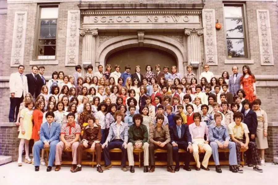 A large group of students and teachers pose for a school photo in front of a brick building labeled "School No Two." Most are wearing colorful 1970s-style clothing.