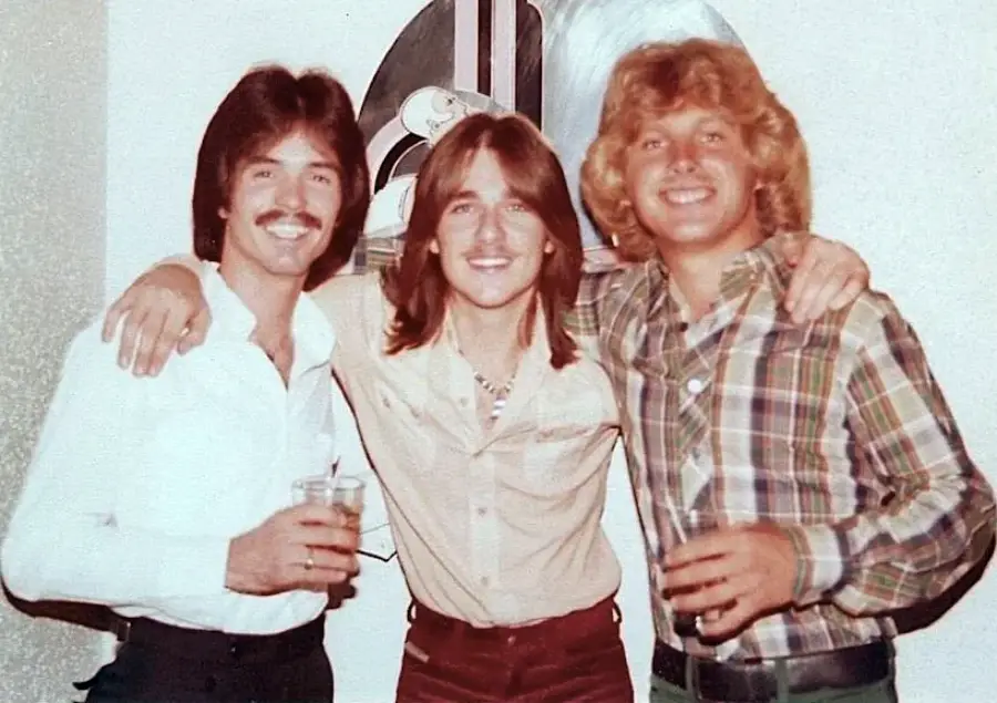 Three young men smiling with arms around each other, dressed in 1970s-style shirts and holding drinks, posing indoors against a light-colored wall with artwork in the background.