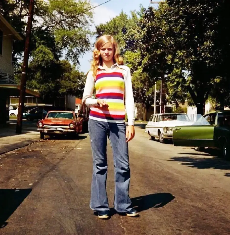 A young person with long blonde hair, wearing a rainbow-striped vest over a white shirt and flared jeans, stands on a sunlit residential street with parked vintage cars and trees in the background.