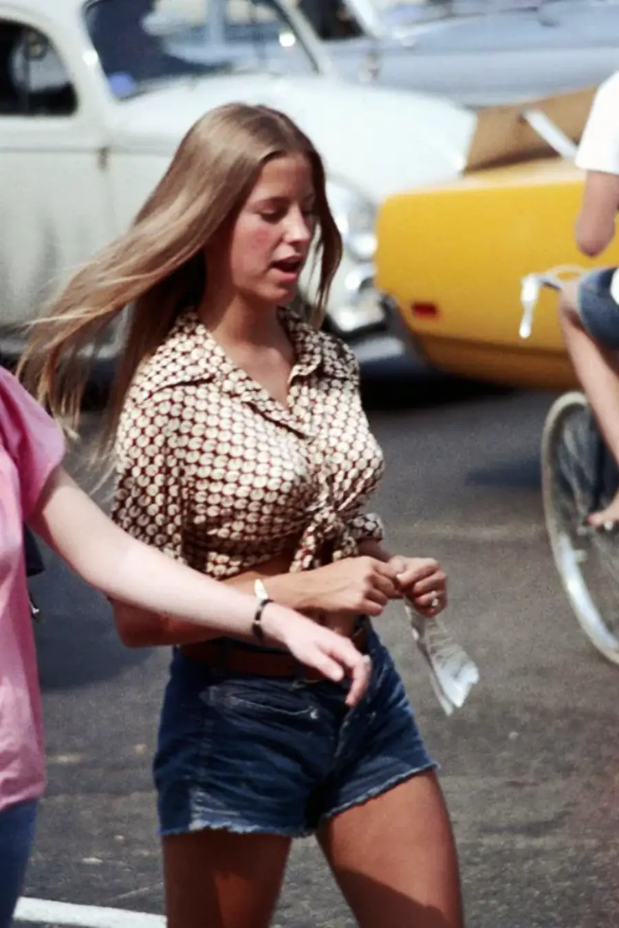 A young woman with long hair wears a patterned, tied-up shirt and denim shorts, walking outdoors. She is holding something in her hand, with cars and a person on a bicycle visible in the background.