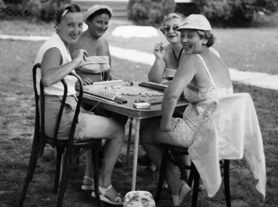 Four women in vintage swimsuits and hats sit around a table outdoors, smiling and playing a board game, possibly mahjong, on a sunny day. Trees and grass are visible in the background.