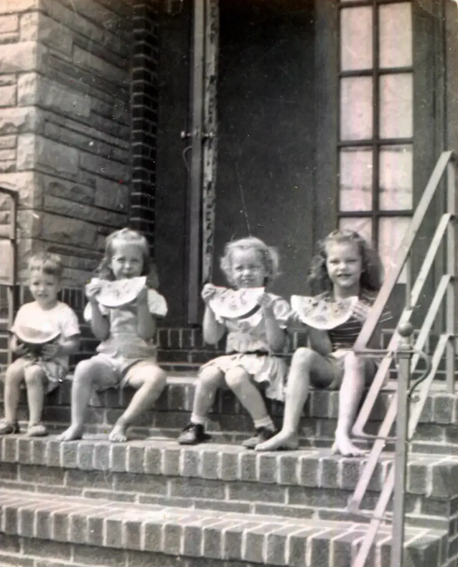 Four young children sit on brick steps outside a building, smiling and holding slices of watermelon. The scene appears to be from an earlier era, with the children dressed in vintage clothing.