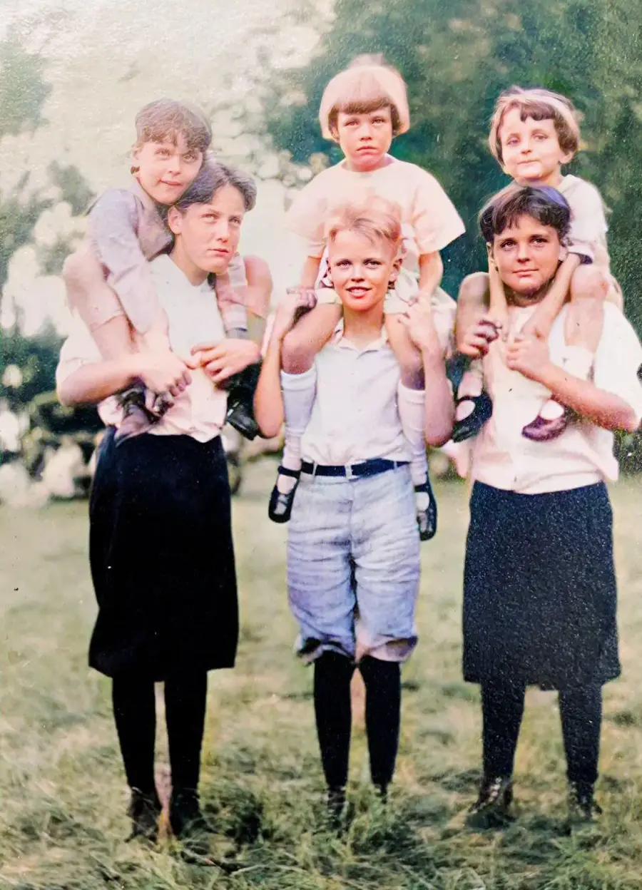 Five children stand outside on grass; three older kids stand in front, each carrying a younger child on their shoulders. All are dressed in old-fashioned clothing, and trees are visible in the blurred background.