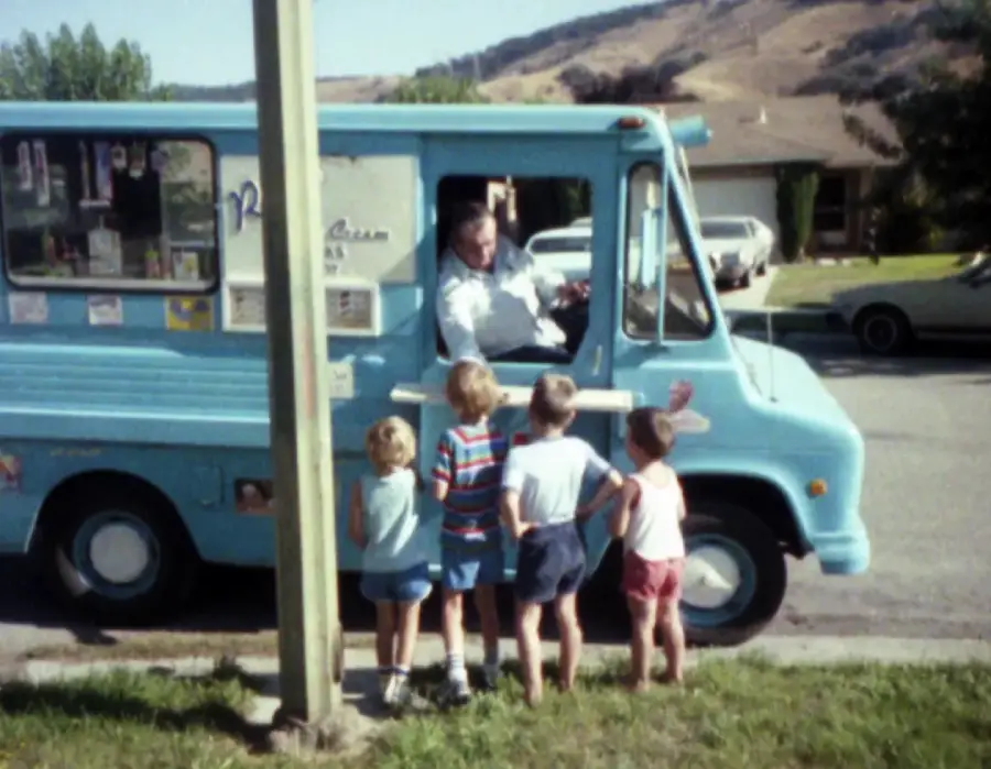 Four young children stand by a blue ice cream truck, talking to the driver through the window on a suburban street. A pole partially blocks the view, and houses and hills are visible in the background.