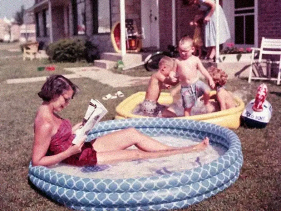 A woman in a red swimsuit relaxes in a small inflatable pool reading a magazine, while three children play in a yellow kiddie pool nearby and another woman stands in the background near a house.