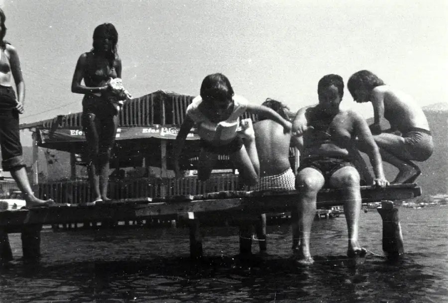 A vintage black-and-white photo of six people, including children and adults, sitting and standing on a wooden dock over water, with a building and mountains visible in the background.