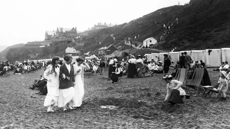 Black and white photo of a crowded beach scene with people in early 20th-century clothing, some sitting on deck chairs, others walking, and buildings visible on a hill in the background.