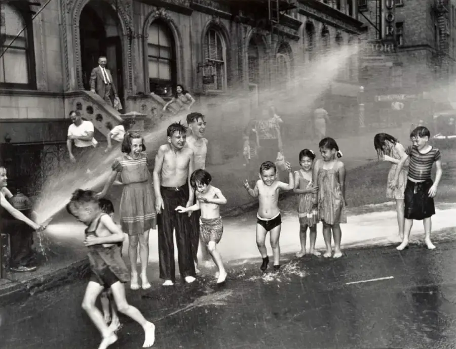 Children and adults laugh and play in the spray of an opened fire hydrant on a city street, with old apartment buildings and steps in the background. Some children are soaked, others dodge the water.