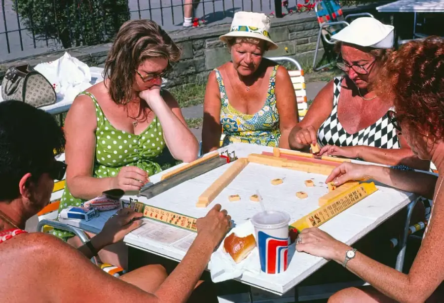 Four women in colorful summer dresses and hats sit around a table outdoors, playing a board game with tiles and racks. A large drink cup and bags are on the table. It is a sunny day.