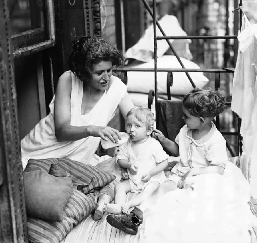 A woman sits on a fire escape with two young children, feeding a baby with a bottle while another child sits beside them. Pillows and blankets are arranged around them in a cozy setting.