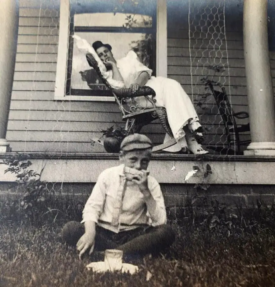 A vintage black-and-white photo shows a woman in a long dress relaxing in a rocking chair on a porch, while a boy in a cap and suspenders sits in the grass below, eating a sandwich and drinking milk.