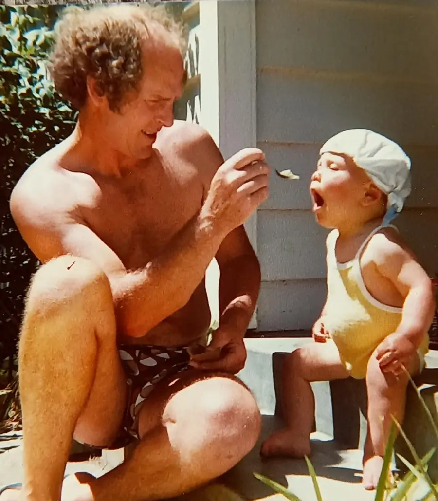 A man in swim trunks sits on outdoor steps, feeding a toddler in a yellow swimsuit and white hat with a spoon. The toddler sits with mouth open, ready for the bite, while sunlight brightens the scene.