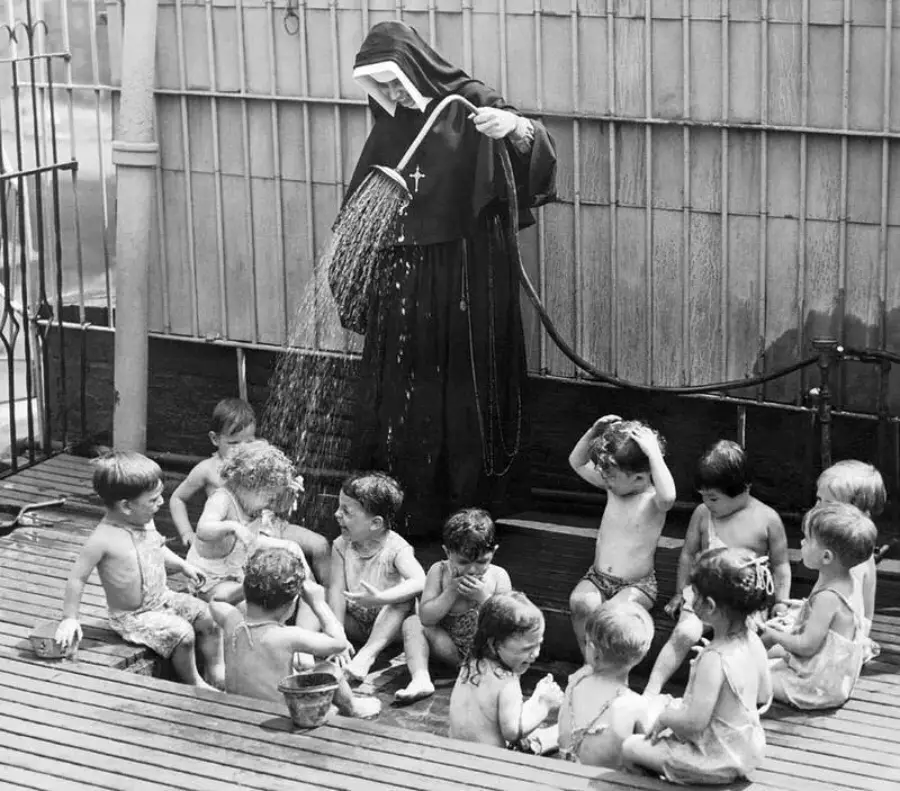 A nun in a habit uses a hose to spray water over a group of young children sitting together outdoors on a wooden deck, while the children laugh, play, and cool off.