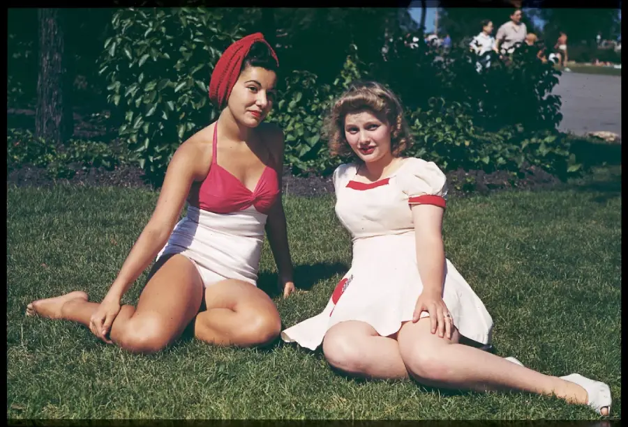 Two women sit on grass in vintage summer outfits; one wears a red headscarf and pink-and-white swimsuit, the other a white dress with red accents. Green bushes and people walking are in the background on a sunny day.