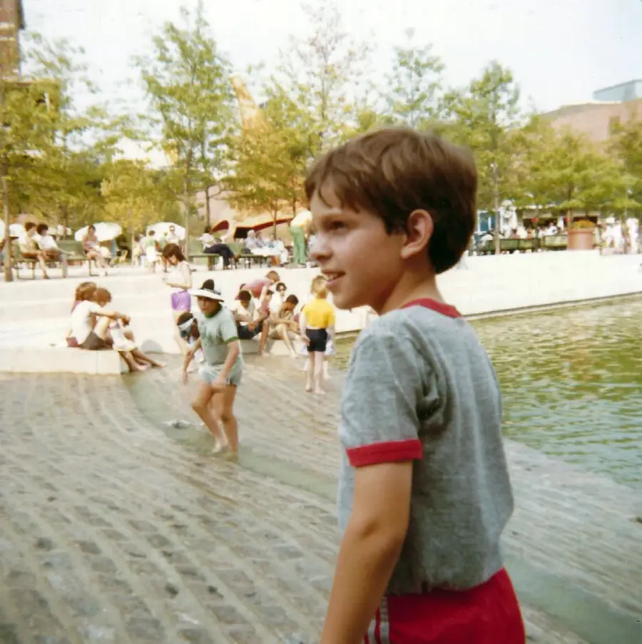 A young boy in a gray shirt with red trim stands near a shallow, stepped fountain. Other children play in the water, while people sit and relax in the background on a sunny day in a public park.