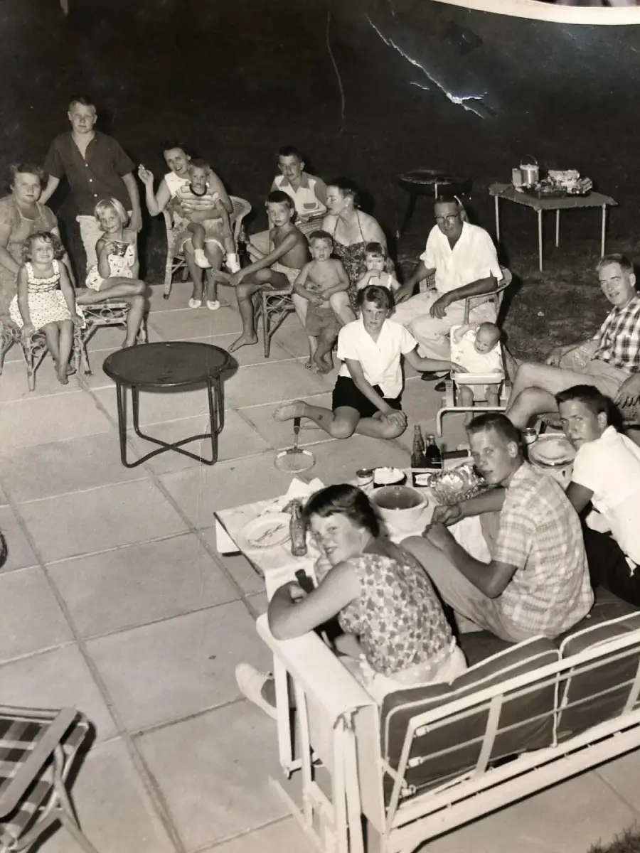 A black-and-white photo shows a large group of adults and children gathered outdoors on a patio. Some sit on chairs and benches, others stand. Food and drinks are on tables. The mood appears lively and relaxed, likely at a family gathering.