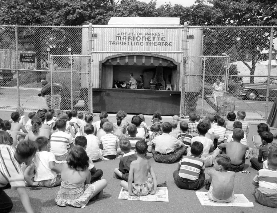 A group of children sit on the ground watching a marionette puppet show in an outdoor theater labeled "Dept. of Parks Marionette Travelling Theatre," surrounded by a chain-link fence.