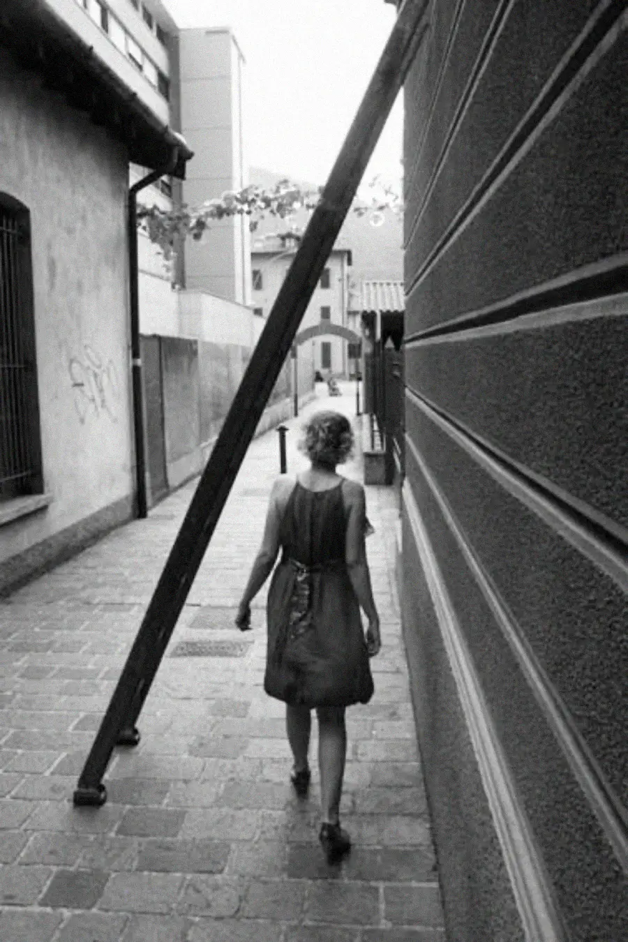 A woman in a dress walks down a narrow cobblestone alley beside a building, passing under a wooden ladder propped diagonally against a wall. The scene is in black and white.
