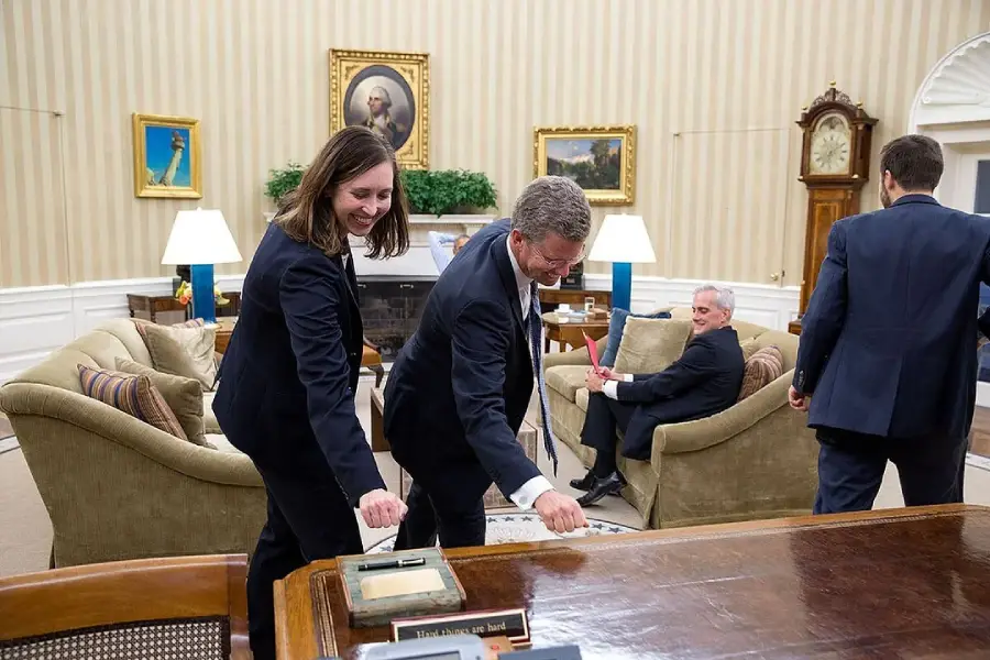 Two people in suits are smiling and pretending to dust or clean a desk in an office with beige walls and classical decor, while two others are in the background, one sitting and another walking away.