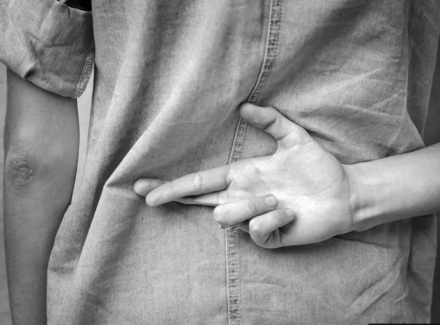 A person stands with their back turned, wearing a button-up shirt, and has their fingers crossed behind their back.