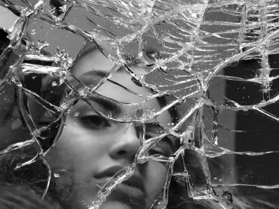 A woman's face is seen through a shattered glass surface, with cracks and fragments partially obscuring her expression. The image is in black and white, creating a dramatic, reflective mood.