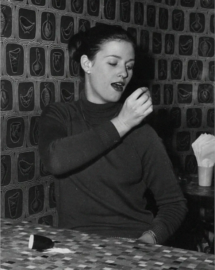 A woman in a turtleneck sweater sits at a mosaic-tiled table, raising her hand to her mouth as if about to eat or taste something. The background features a patterned wall with abstract designs.