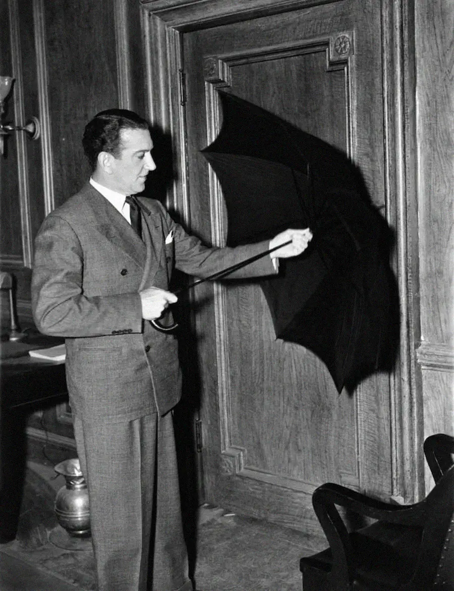 A man in a suit stands indoors, holding an open umbrella and pointing it toward a closed wooden door. The room features wooden paneling and classic furnishings.